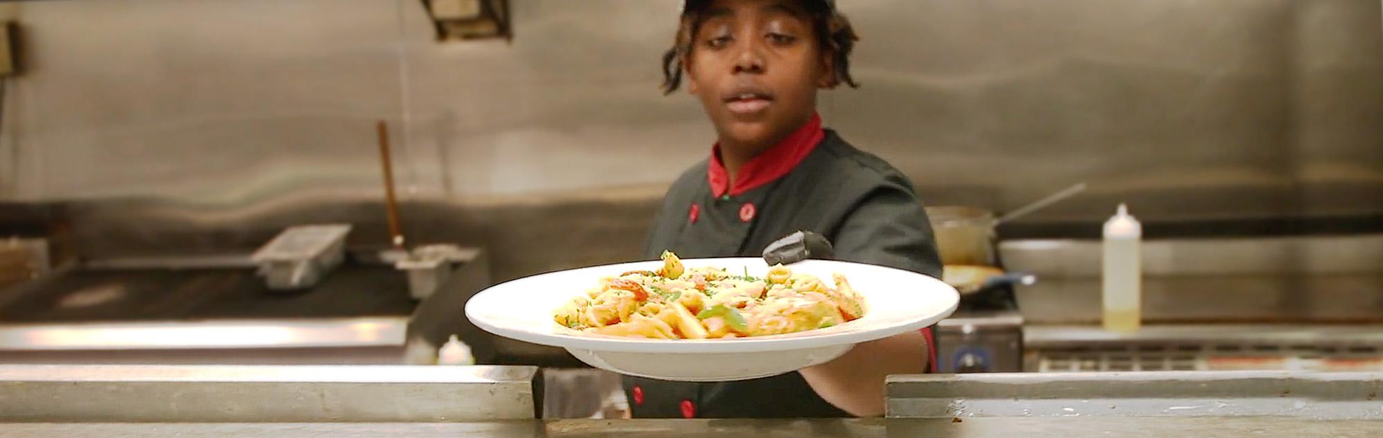 Chef hands plate of pasta from kitchen to server.